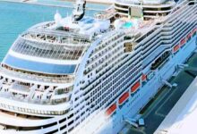 O navio da MSC Cruzeiros ancorado em um porto de águas cristalinas, cercado por montanhas e um céu azul intenso. Atrás do navio, outros barcos e edifícios do porto podem ser vistos