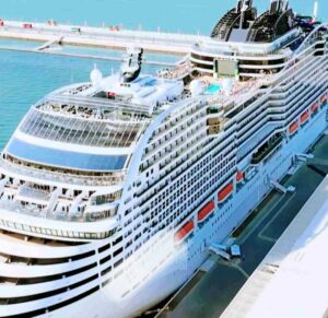 O navio da MSC Cruzeiros ancorado em um porto de águas cristalinas, cercado por montanhas e um céu azul intenso. Atrás do navio, outros barcos e edifícios do porto podem ser vistos