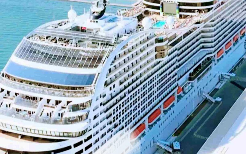 O navio da MSC Cruzeiros ancorado em um porto de águas cristalinas, cercado por montanhas e um céu azul intenso. Atrás do navio, outros barcos e edifícios do porto podem ser vistos