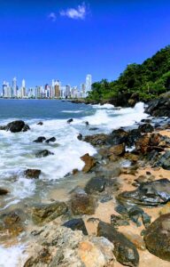 Explore a deslumbrante paisagem de Balneário Camboriú. Uma visão serena do mar azul, a vegetação exuberante à beira-mar, a vibrante cidade à distância e a praia com sua areia dourada e pedras pitorescas. Um cenário encantador que convida você a desfrutar de momentos de tranquilidade e contemplação.