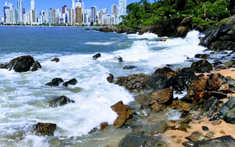 Explore a deslumbrante paisagem de Balneário Camboriú. Uma visão serena do mar azul, a vegetação exuberante à beira-mar, a vibrante cidade à distância e a praia com sua areia dourada e pedras pitorescas. Um cenário encantador que convida você a desfrutar de momentos de tranquilidade e contemplação.